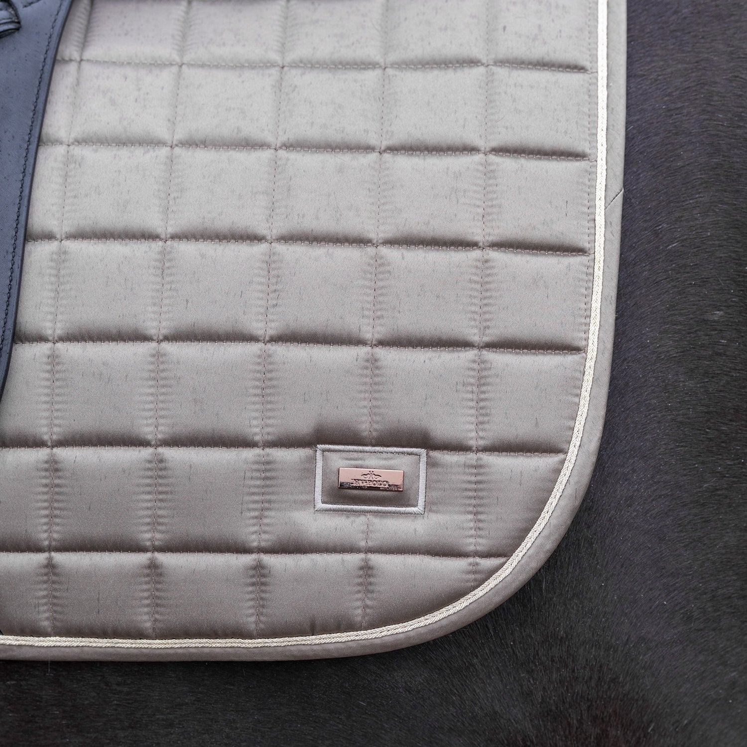 Die HV Polo Schabracke HVPDewi in der Farbe stone in einem schönen Beigeton hat abgerundete Linien. Unter dem Sattel auf einem dunklen Pferd wirkt die Schabracke sehr elegant. Das rose goldene metallische HV Polo Logo ist klein und dennoch edel. Die goldene Kordel ist hell und umfährt die Schabracke wie eine kontrastfarbige Linie.