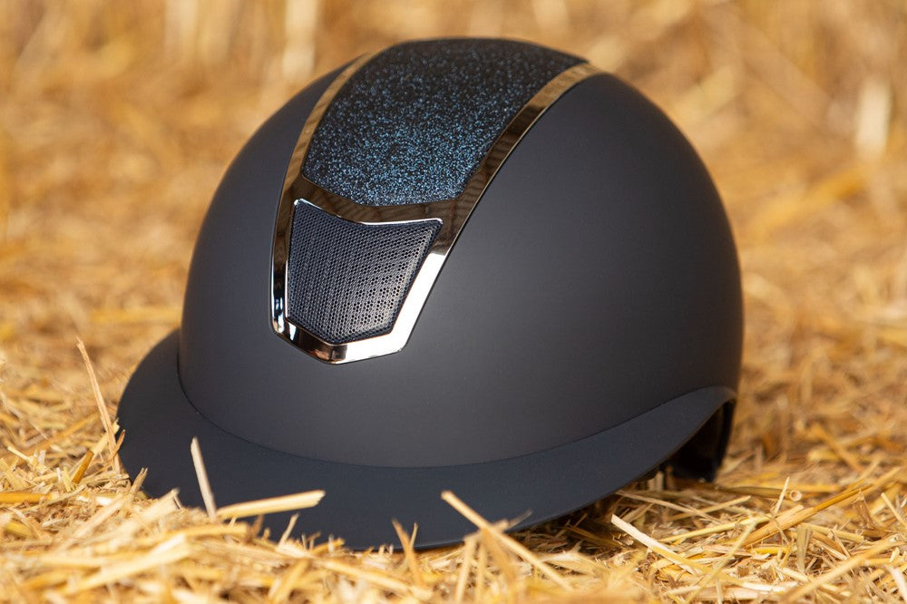 Das Lifestylebild zeigt den Harry’s Horse Sicherheitsreithelm Mont Blanc Sparkle in navy silber auf einem Heuballen liegend, wobei die besondere Farbgebung und der funkelnde Glitzer im Frontbereich deutlich erkennbar sind, was den eleganten Reithelm ideal für Turnierreiter und Freizeitreiter macht, die modisches Design mit hoher Sicherheit und zuverlässigem Schutz bei Stürzen kombinieren möchten.