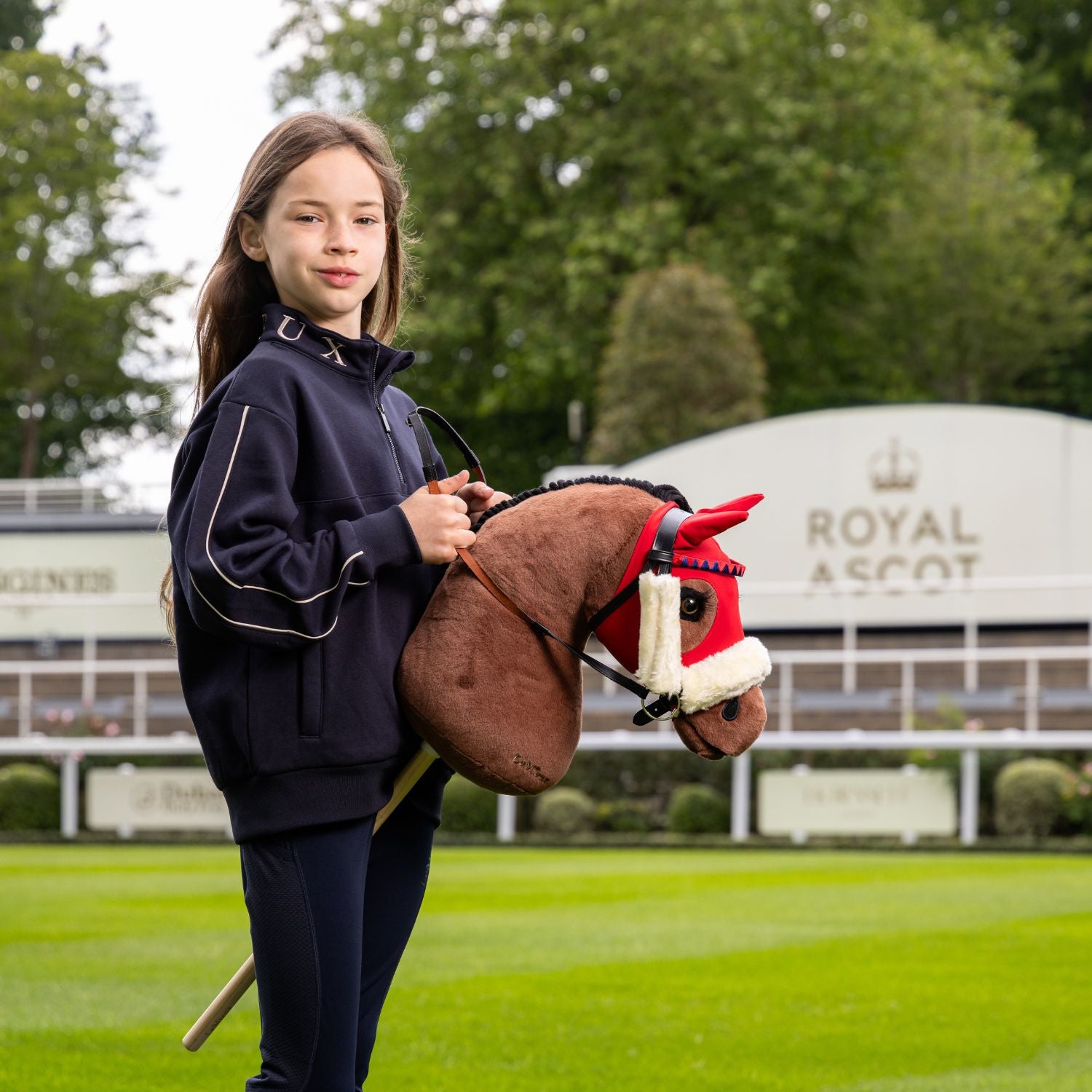 Braunes Steckenpferd Chancer mit weißer Blesse und Holzstab wird von junger Reiterin geritten. Im Hintergrund die Rennbahn Royal Ascot. Das Hobby Horse trägt eine Renntrense und Rennmaske wie ein echter Vollblüter auf der Rennbahn.