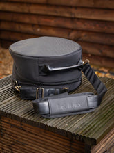 LeMieux Helmtasche Luxe Schwarz liegt flach vor dem Stall mit Schultergurt daneben – edle, gepolsterte Reithelm Tasche mit goldenen Details.