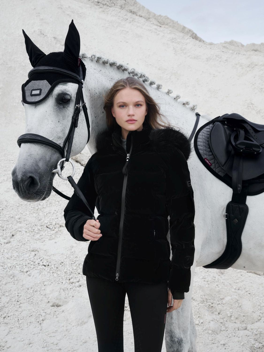 Reiterin trägt die LeMieux Aspen Puffer Jacket Black beim Führen des Pferdes – stilvolle und warme Reitjacke in Samtoptik. Die Aspen Steppjacke kann mit allen Teilen der Aspen Capsual Kollektion kombiniert werden. Hier kombiniert mit der Aspen Herringbone Fliegenmütze, Schabracke und schwarzer Lemieux Reithose.