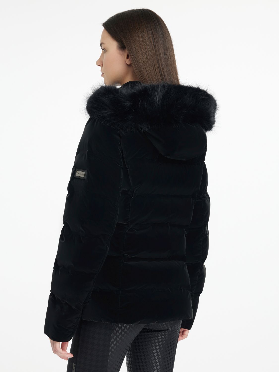 Rückansicht der LeMieux Aspen Puffer Jacket Black – taillierte schwarze Steppjacke mit abnehmbarer Kapuze. Model trägt die Steppjacke Aspen und zeigt diese in seitlicher Rückansicht. LeMieux Logo am Arm ist zu erkennen.