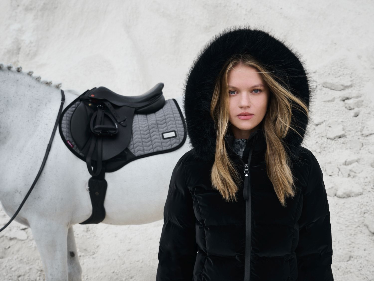 Modelt trägt die LeMieux Steppjacke Aspen mit hochgezogener Kapuze. Der Fellrand schützt vor kaltem Wind. Im Hintergrund steht ein gesatteltes Pferd mit der Springschabracke Aspen Close Contact Square Herringbone.