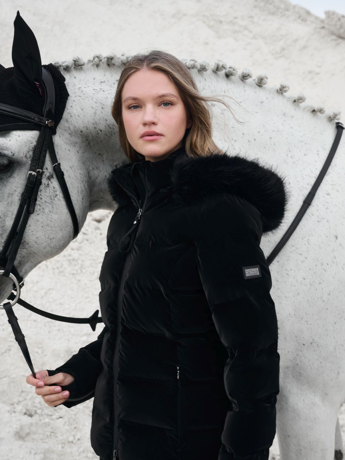 Die Samtoptik in schwarz steht zum Kontrast zum weißen Pferd im Hintergrund. Reiterin zeigt die Aspen Jacke seitlich, sodass das Lemieux Logo am linken Arm zu erkennen ist. Die Kapuze hängt runter, Fellrand wirkt voluminös.