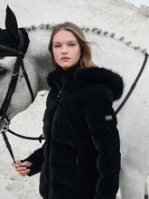 Die Samtoptik in schwarz steht zum Kontrast zum weißen Pferd im Hintergrund. Reiterin zeigt die Aspen Jacke seitlich, sodass das Lemieux Logo am linken Arm zu erkennen ist. Die Kapuze hängt runter, Fellrand wirkt voluminös.
