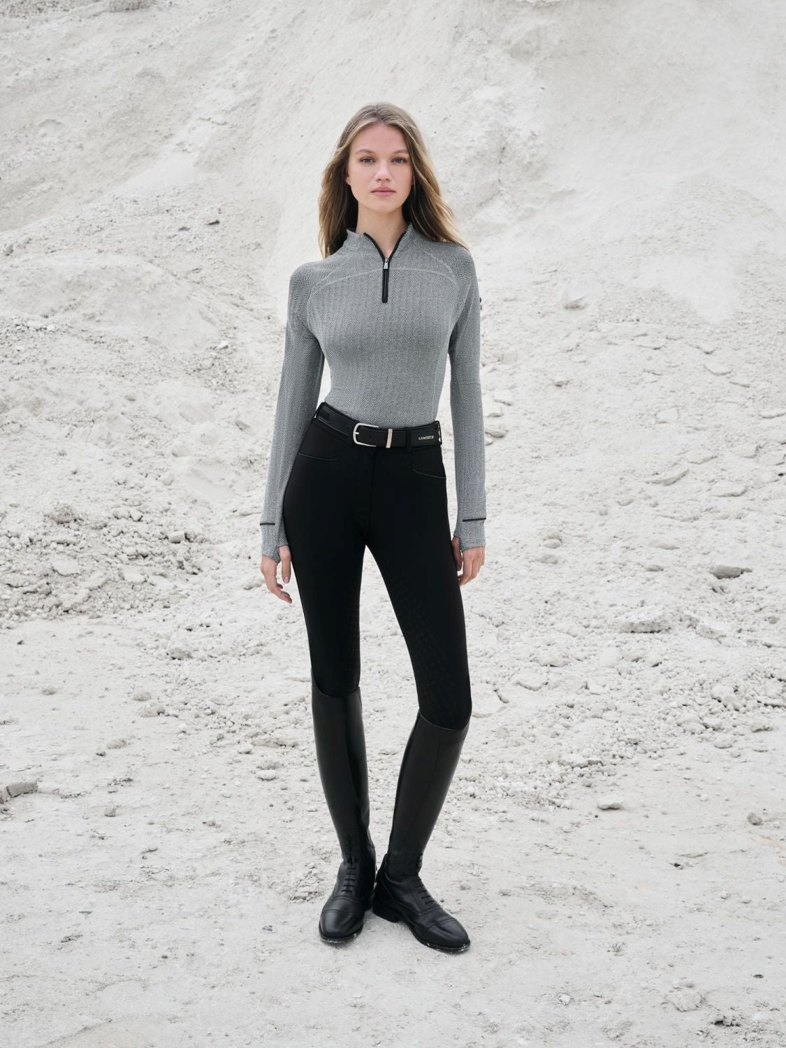 Reitmode von LeMieux – Model zeigt den schwarzen Aspen Herringbone Base Layer mit elegantem Fischgrät-Design. Halb offener Stehkragen mit Reißverschluss. Kombiniert mit schwarzer Reithose und Stiefeln. Helle und Dunkle Farben lassen sich zur Aspen Kollektion sehr gut kombinieren. Die Kapsel-Kollektion ist vielseitig nutzbar und mit anderen Kleidungsteilen von LeMieux kombinierbar.