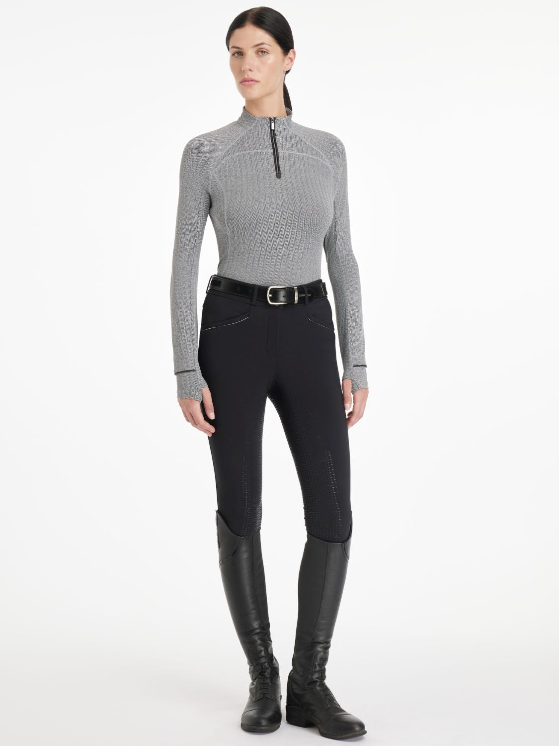 Reiterin kombiniert den LeMieux Aspen Herringbone Base Layer Black mit einer schwarzen LeMieux Reithose, Gürtel und Reitstiefel. Dadurch wirkt das edle Fischgrätmuster wie ein zeitloses Design. An den Ärmeln sind die Daumenlöcher zu erkennen, die das sportliche Langarmshirt sehr modern wirken lassen.