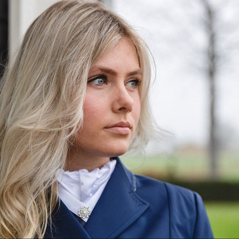 Eine Dressurreiterin trägt das elegante ANKY Plastron Symphony in Weiß unter einem dunkelblauen, sehr hoch geschlossenen Reitjacket. Diese Aufnahme demonstriert, dass das Turnierplastron ideal auch für hohe Kragenabschlüsse oder einen Dressurfrack geeignet ist. Trotz des geschlossenen Schnitts ragen der feine, plissierte Kragen mit seinen Glitzer-Kristallsteinen und die funkelnde, sternenförmige Plastronnadel bestechend schön und gut sichtbar aus dem Ausschnitt hervor.