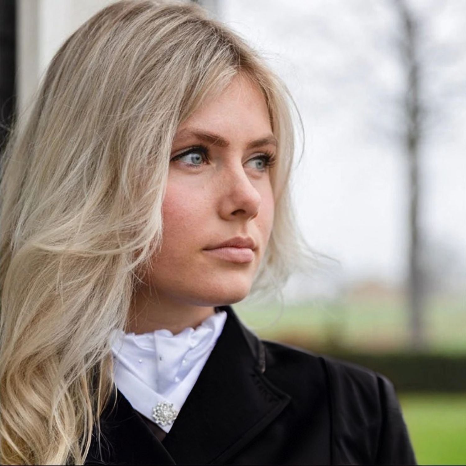 Eine Dressurreiterin trägt das elegante ANKY Plastron Symphony in Weiß unter einem dSchwarzen, sehr hoch geschlossenen Reitjacket. Diese Aufnahme demonstriert, dass das Turnierplastron ideal auch für hohe Kragenabschlüsse oder einen Dressurfrack geeignet ist. Trotz des geschlossenen Schnitts ragen der feine, plissierte Kragen mit seinen Glitzer-Kristallsteinen und die funkelnde, sternenförmige Plastronnadel bestechend schön und gut sichtbar aus dem Ausschnitt hervor.