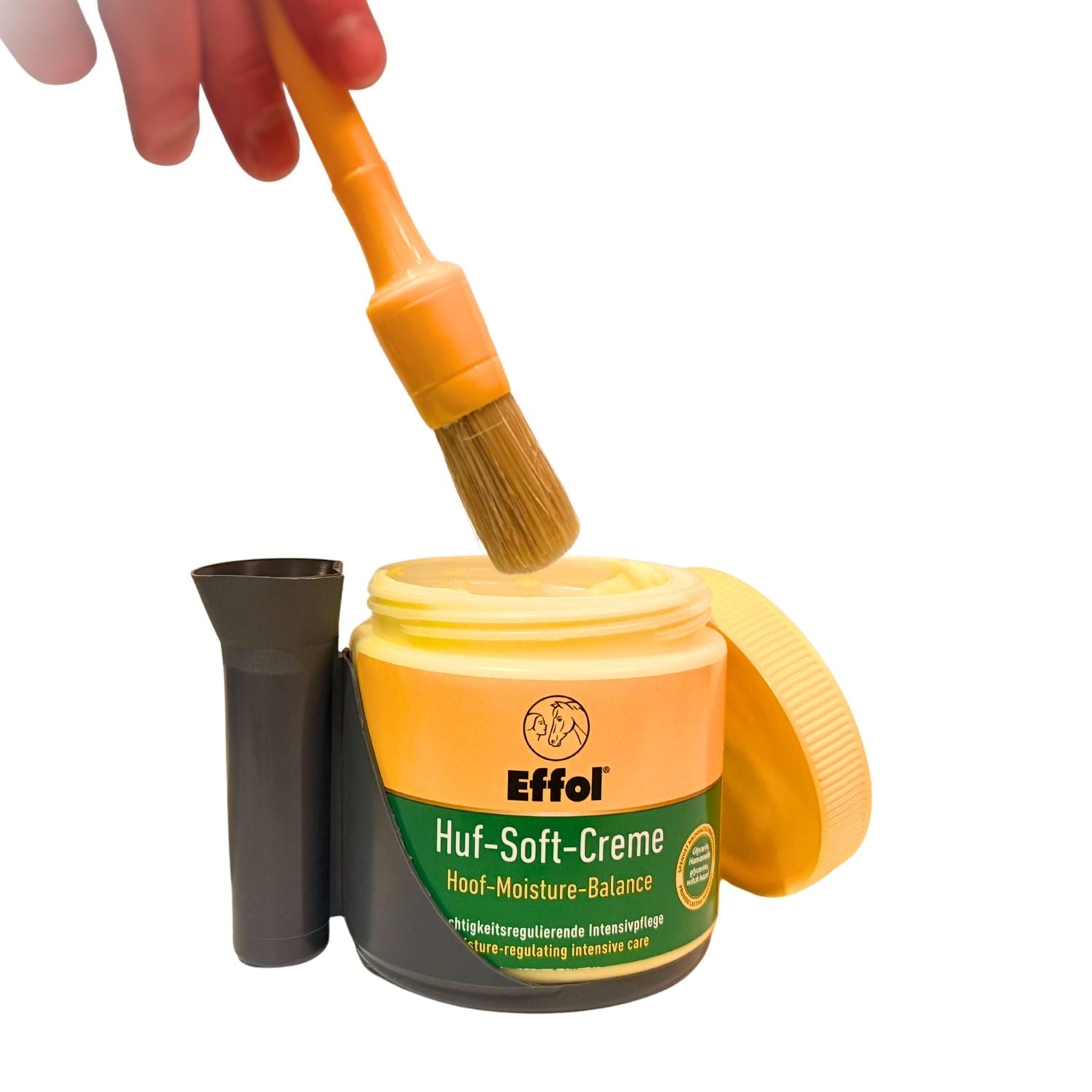 Das Bild zeigt den Effol Pinsel-Fix bei der Hufpflege mit einer geöffneten Effol Huf-Soft-Creme auf der Standplattform, während eine Hand den Pinsel in die Creme eintaucht und die andere Hand frei bleibt, um den Pferdehuf sicher zu halten, wodurch eine hygienische und saubere einhändige Anwendung ermöglicht wird.