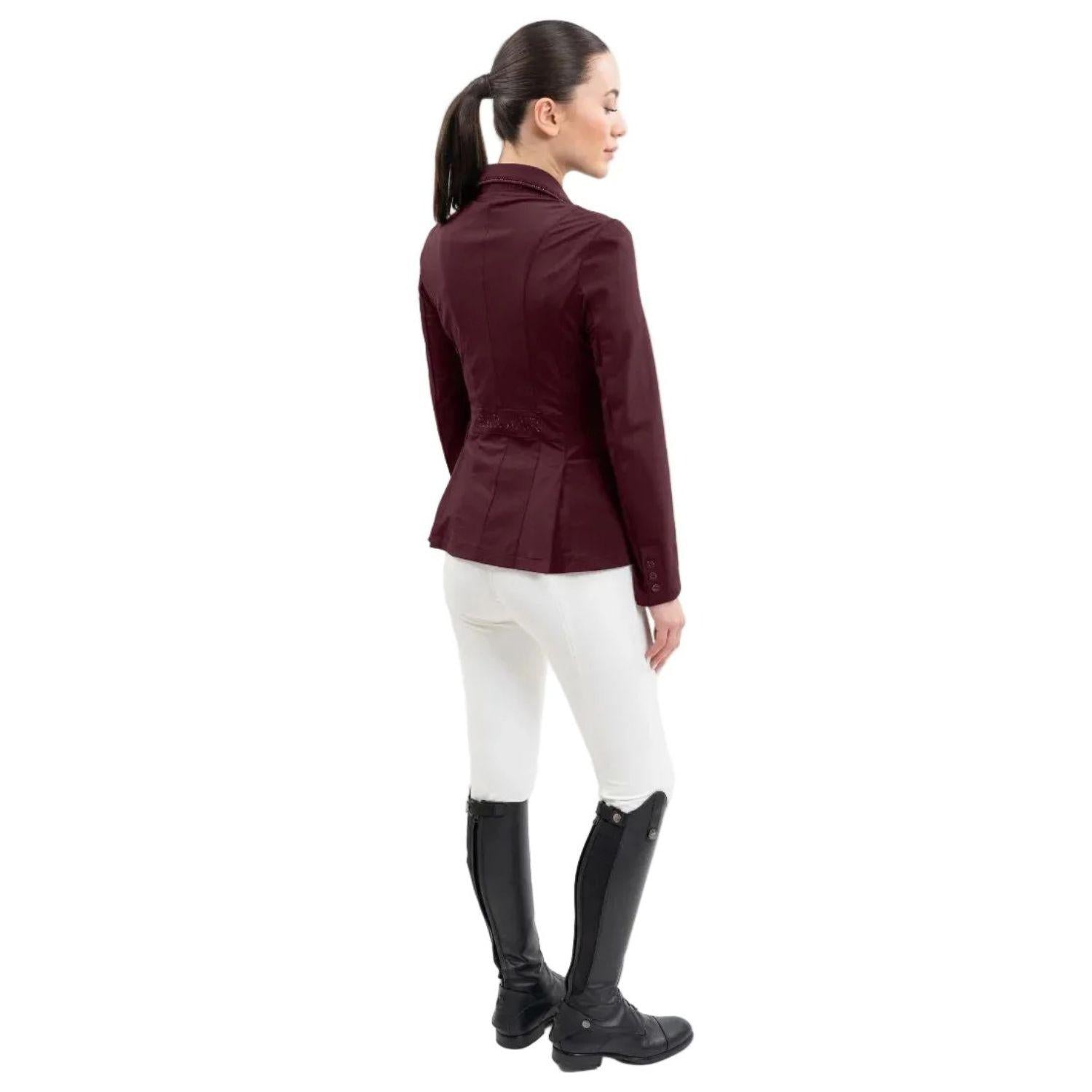 Rückansicht einer Reiterin im Harcour Damen Turnierjacket Oria. Die luxuriöse Farbe Aubergine, ein dunkler Rotton mit kühlen Violett- und warmen Brauntönen, harmoniert perfekt mit weißer Turnierkleidung. An der Taille funkelt der edle Harcour Glitzerschriftzug. Das elegante Schößchen, oft auch Reitrock genannt, bietet dank zwei seitlichen Schlitzen höchste Flexibilität und Bewegungsfreiheit im Sattel. Ein exklusives Reitsakko, das im Turniersport durch edles Design perfekt in Szene gesetzt wird.