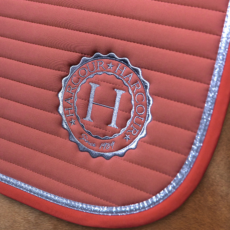 Gestochen scharfes Detailbild der linken unteren Seite der Harcour Schabracke Karembar in der Farbe Aurora Pink. Die Aufnahme zeigt die präzise horizontale Steppung, das farblich abgesetzte, dunklere rosa Einfassband und die flache, silberne glitzernde Litze. Diese funkelnde Litze passt perfekt zu dem edlen, runden, silber eingestickten Harcour Logo auf der Sattelunterlage. Ein echter stylischer Blickfang und Garant für einen perfekten Auftritt, egal ob im Dressursattel, Springsattel oder Ponysattel.