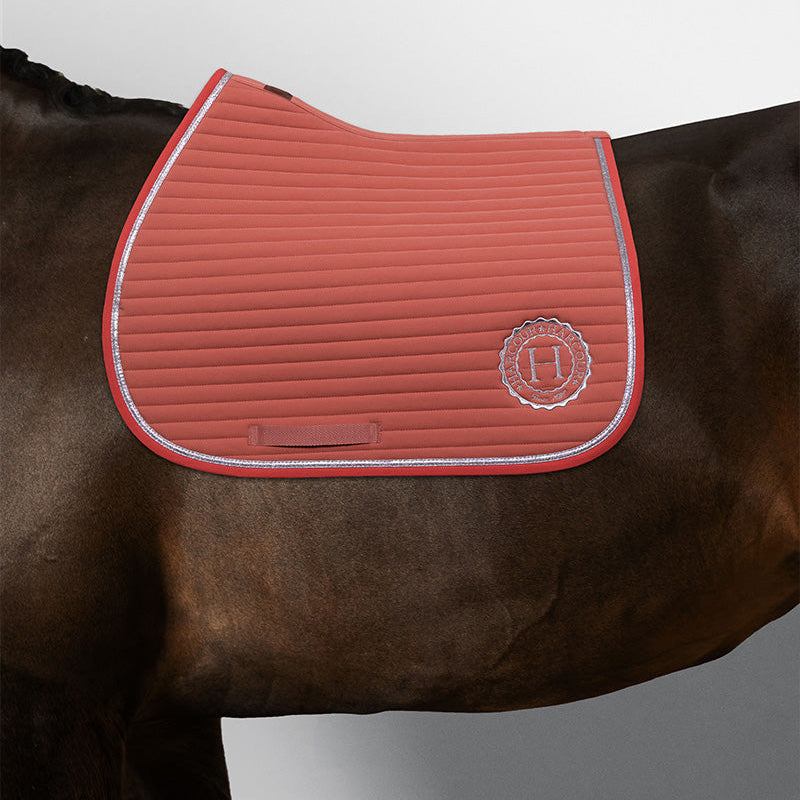 Harcour Schabracke Karembar in Aurora Pink auf einem dunkelbraunen Pferd. Diese anatomische Springschabracke liegt optimal unter gängigen Springsätteln und ist der robuste, aber elegante Begleiter für das Springtraining und ausgedehnte Ausritte. Details: große Widerristfreiheit, die horizontale Steppung und die silberglänzende Litze im Fokus. Ausdrucksstarker Kontrast: Das frische Rosa leuchtet intensiv und warm auf dem dunklen Fell. Eine sportlich-elegante Sattelunterlage für ambitionierte Reiterinnen.