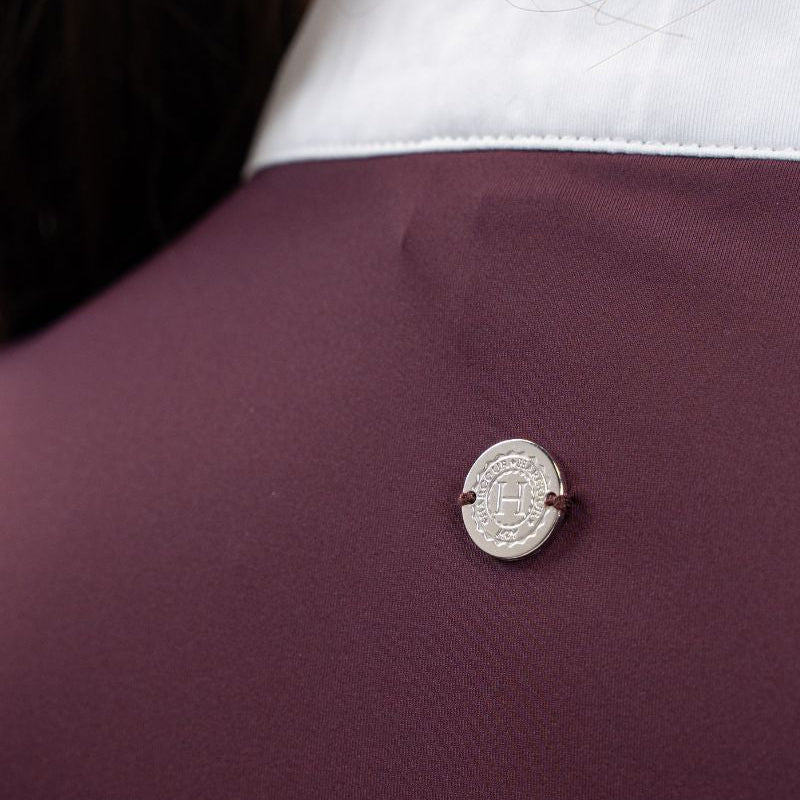 Die Rückansicht des Harcour-Turniershirts Shine aubergine, einem dunklen Violettton, mit dem dezenten, runden, silbernen Logo auf dem elastischen Stretchmaterial für optimale Bewegungsfreiheit. Der darüber liegende Stehkragen ist kontrastfarbig in Weiß abgesetzt.