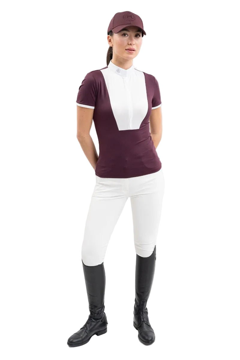 Eine Turnierreiterin im kompletten, eleganten Turnieroutfit trägt frontal das violette Harcour Kurzarm-Turniershirt Shine Aubergine mit edlem weißem Latz, passendem Stehkragen und kontrastfarbigen Ärmelabschlüssen, kombiniert mit einer weißen Harcour Reithose Jaltika und schwarzen Lederreitstiefeln für ein harmonisches Erscheinungsbild im Dressursport wie Springsport an warmen Sommertagen.