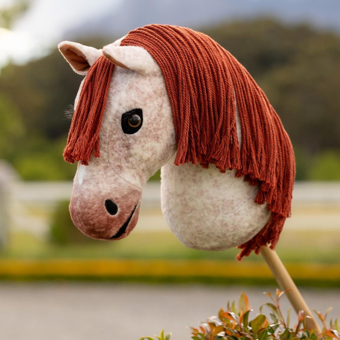 LeMieux Hobby Horse Poppy draußen vor einem Reitplatz. Das Rotschimmel-Fell zeigt leichte Rotverläufe im Kontrast zur roten Mähne und dem plüschigen Maul