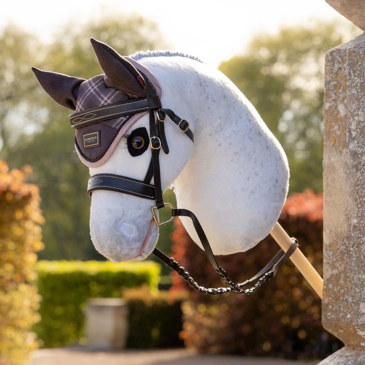 Detailaufnahme des LeMieux Hobby Horse Coco: Der weiße Schimmelkopf im Profil an einem Steinpfeiler, ausgestattet mit der braunen Leder-Trense Hunter und karierter Heritage Fliegenhaube in Cocoa für anspruchsvolle Hobby Horsing Turniere.