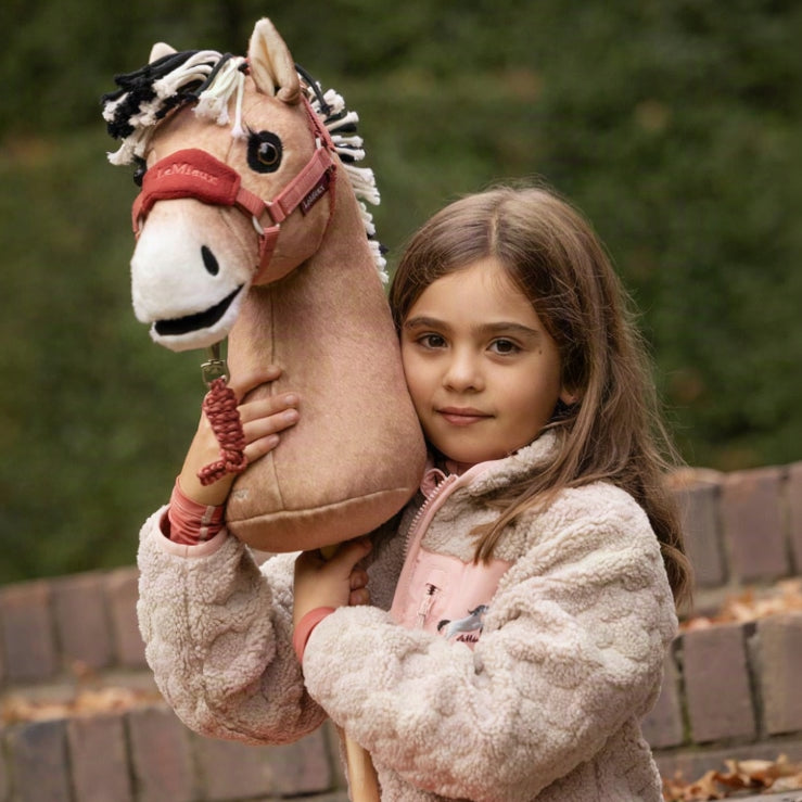Nahaufnahme: Mädchen hält das LeMieux Hobby Horse Oskar (norwegisches Fjordpferd als Steckenpferd) im Arm. Das Falbe-Pony mit zweifarbiger Stehmähne, Mehlmaul und bernsteinfarbenen Augen trägt das LeMieux Hobby-Horse-Halfter mit Führstrick in Spiced Earth.