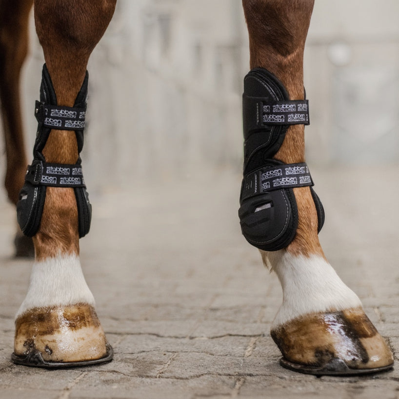 Dieses Bild zeigt die schwarzen Stübben Hybrid Springgamaschen im Einsatz an den Vorderbeinen eines Springpferdes, wo sie das Pferdebein im Bereich der Sehnen sicher umschließen. Der auffällige Stübben Evolution Allover-Print an den elastischen Verschlüssen setzt einen optischen Akzent, während die robuste Hartschale zuverlässig vor Verletzungen durch Eisen und Hufe der Hinterbeine beim Springen oder im intensiven Training schützt.