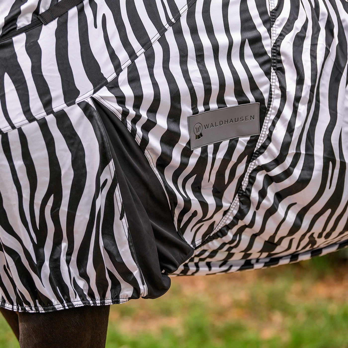 Detailaufnahme der Waldhausen Ekzemerdecke Zebra Premium in schwarz-weiß, die die dunkel abgesetzte Gehfalte und das sichtbare Waldhausen-Logo zeigt. Dieses Zebra-Design bietet Pferden und Ponys maximale Bewegungsfreiheit auf der Weide beim Galoppieren, Spielen und Buckeln. Der funktionale Zebra-Print dient als mechanischer Fliegenschutz zur Abwehr von Kriebelmücken, während die elastische Gehfalte dieser Sommerdecke eine optimale, körpernahe Passform beim Freilauf.