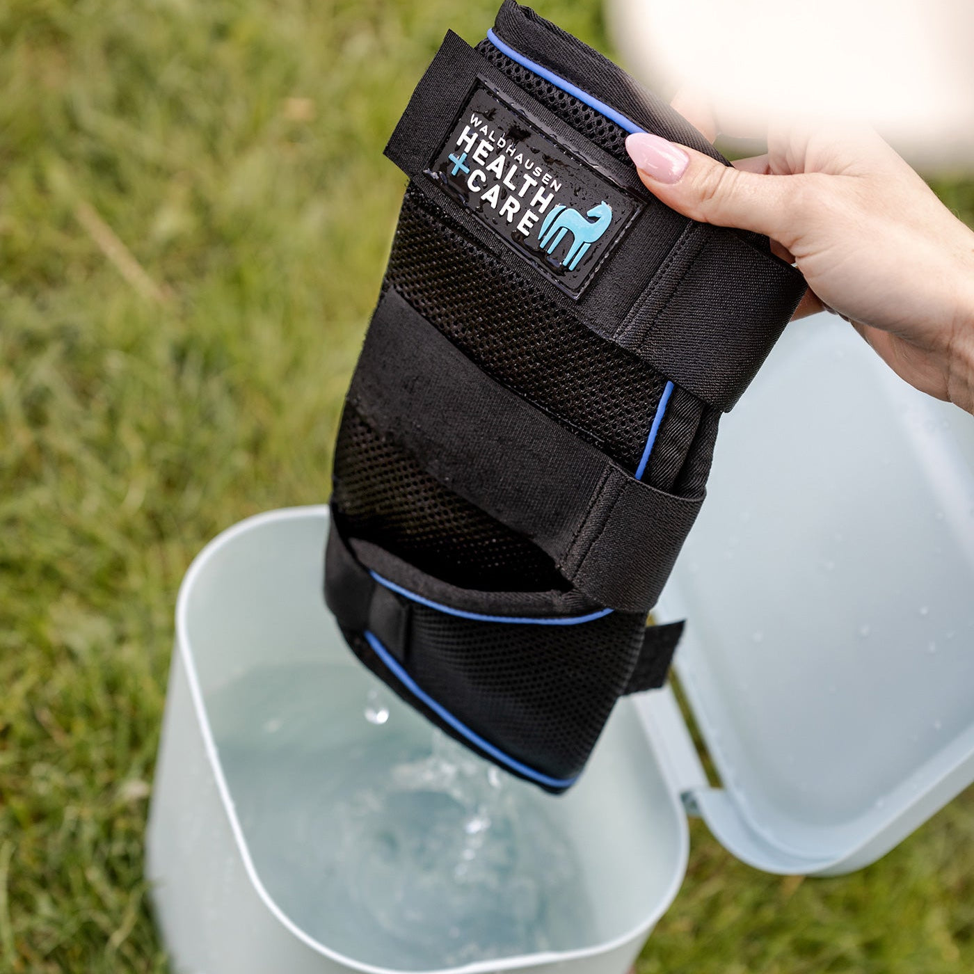 Eine Reiterin nimmt die nasse, mit Wasser vollgesaugte Waldhausen Health + Care Ice-Layer Kühlgamasche aus der Waldhausen Multibag ECO Tasche heraus. Gezeigt wird das leichte Auswringen der Gamasche vor dem Anlegen an das Pferdebein. Die formstabile Kühlgamasche wird anschließend per Klettverschluss fixiert, um durch natürliche Verdunstungskälte einen langanhaltenden Cooling-Effekt zu erzielen. Perfekt für die mobile Pferdepflege im Sommer.