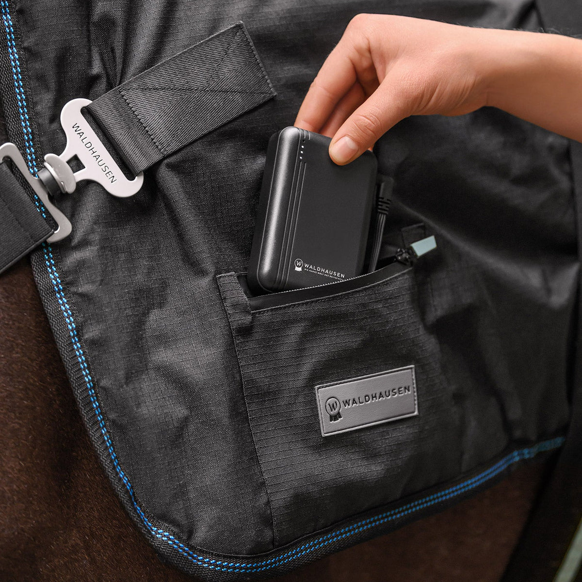 Nahaufnahme der Handhabung an der Waldhausen Health + Care TecSupreme Decke: Eine Hand setzt die kompakte Waldhausen-Powerbank mit USB Anschluss in die integrierte Reißverschlusstasche am Pferd ein, um die Heiz- und Massagefunktion kabellos und flexibel mit Strom zu versorgen.