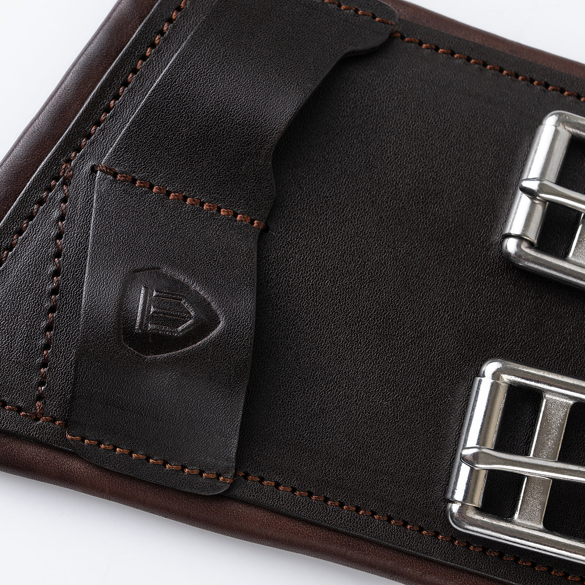 Detailansicht der Leder-Lasche mit geprägtem Winderen-Logo am braunen Dressursattelgurt aus Vollnarbenleder. Die flache Lederführung sorgt für ein ordentliches Anliegen der Gurtstrippen oberhalb der beiden Edelstahl-Rollenschnallen und verhindert Druckstellen am Pferdebauch.