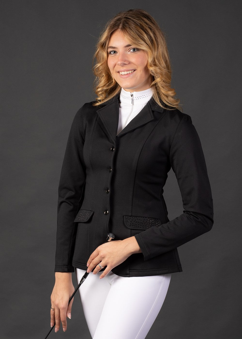 Reiterin trägt das Harry’s Horse Turnierjacket Crystal in Schwarz zusammen mit weißer Turnierreithose und einem passenden Turniershirt. Der kurze, taillierte Schnitt des Jackets bietet maximale Bewegungsfreiheit und eignet sich ideal für Turnierreiterinnen, Reitprofis und Reitabzeichenprüfungen. Das Outfit vereint Stil, Komfort und klassische Turnieroptik.