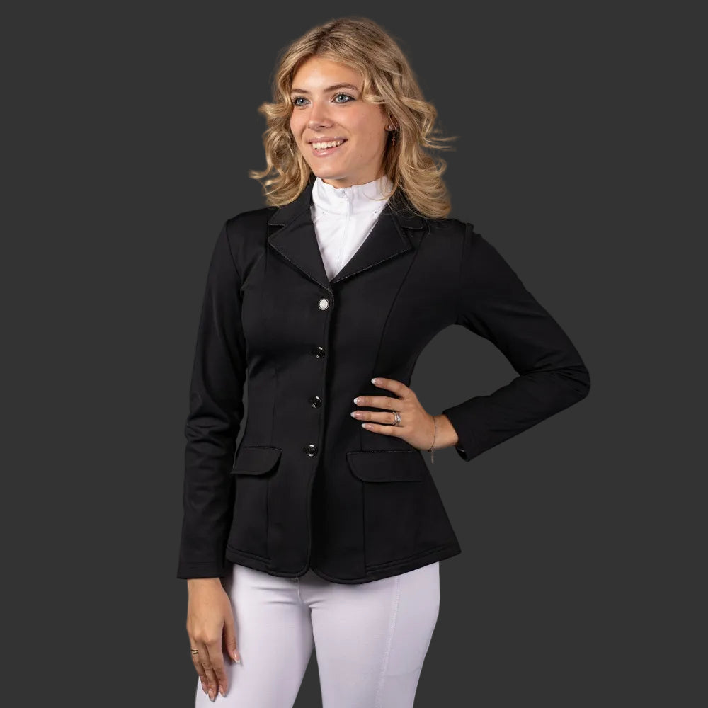 Eine Reiterin trägt das schwarze Turnierjacket „Sparkle“ von Harrys Horse in Schwarz mit Glitzerpaspelierung. Das Turnierjacket ist mit einem weißen Turniershirt und einer weißen Turnierreithose kombiniert. Das Turniersakko ist tailliert und mit einer dezenten Glitzerpaspelierung an den Taschen und dem Kragen versehen. Diese Details veredeln die Turnierreitbekleidung für Damen.