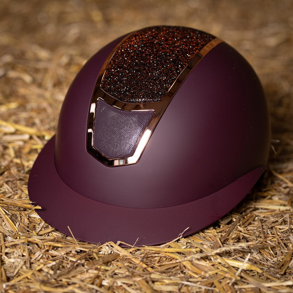 Das Lifestylebild zeigt den Harry’s Horse Sicherheitsreithelm Mont Blanc Sparkle in bordeaux rosegold auf einem Heuballen liegend, wobei die besondere Farbgebung und der funkelnde Glitzer im Frontbereich deutlich erkennbar sind, was den eleganten Reithelm ideal für Turnierreiter und Freizeitreiter macht, die modisches Design mit hoher Sicherheit und zuverlässigem Schutz bei Stürzen kombinieren möchten.