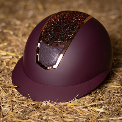 Das Lifestylebild zeigt den Harry’s Horse Sicherheitsreithelm Mont Blanc Sparkle in bordeaux rosegold auf einem Heuballen liegend, wobei die besondere Farbgebung und der funkelnde Glitzer im Frontbereich deutlich erkennbar sind, was den eleganten Reithelm ideal für Turnierreiter und Freizeitreiter macht, die modisches Design mit hoher Sicherheit und zuverlässigem Schutz bei Stürzen kombinieren möchten.