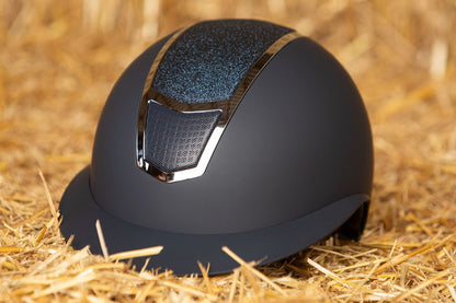 Das Lifestylebild zeigt den Harry’s Horse Sicherheitsreithelm Mont Blanc Sparkle in navy silber auf einem Heuballen liegend, wobei die besondere Farbgebung und der funkelnde Glitzer im Frontbereich deutlich erkennbar sind, was den eleganten Reithelm ideal für Turnierreiter und Freizeitreiter macht, die modisches Design mit hoher Sicherheit und zuverlässigem Schutz bei Stürzen kombinieren möchten.