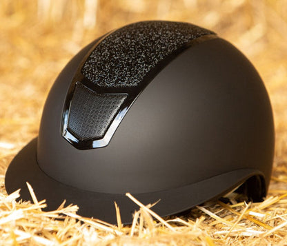 Das Lifestylebild zeigt den Harry’s Horse Sicherheitsreithelm Mont Blanc Sparkle in schwarz-schwarz auf einem Heuballen liegend, wobei die besondere Farbgebung und der funkelnde Glitzer im Frontbereich deutlich erkennbar sind, was den eleganten Reithelm ideal für Turnierreiter und Freizeitreiter macht, die modisches Design mit hoher Sicherheit und zuverlässigem Schutz bei Stürzen kombinieren möchten.