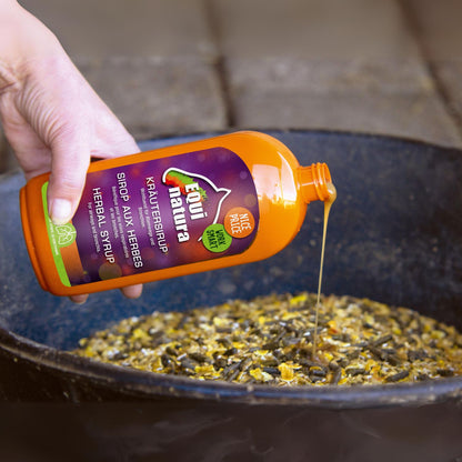 Equinatura Kräutersirup 500 ml – Ergänzungsfuttermittel für Pferde. Eine Hand gießt den pflanzlichen Sirup aus einer orangefarbenen Flasche mit lila Etikett über Pferdefutter in einer Schüssel. Enthält Kräuter zur Unterstützung der Atemwege.