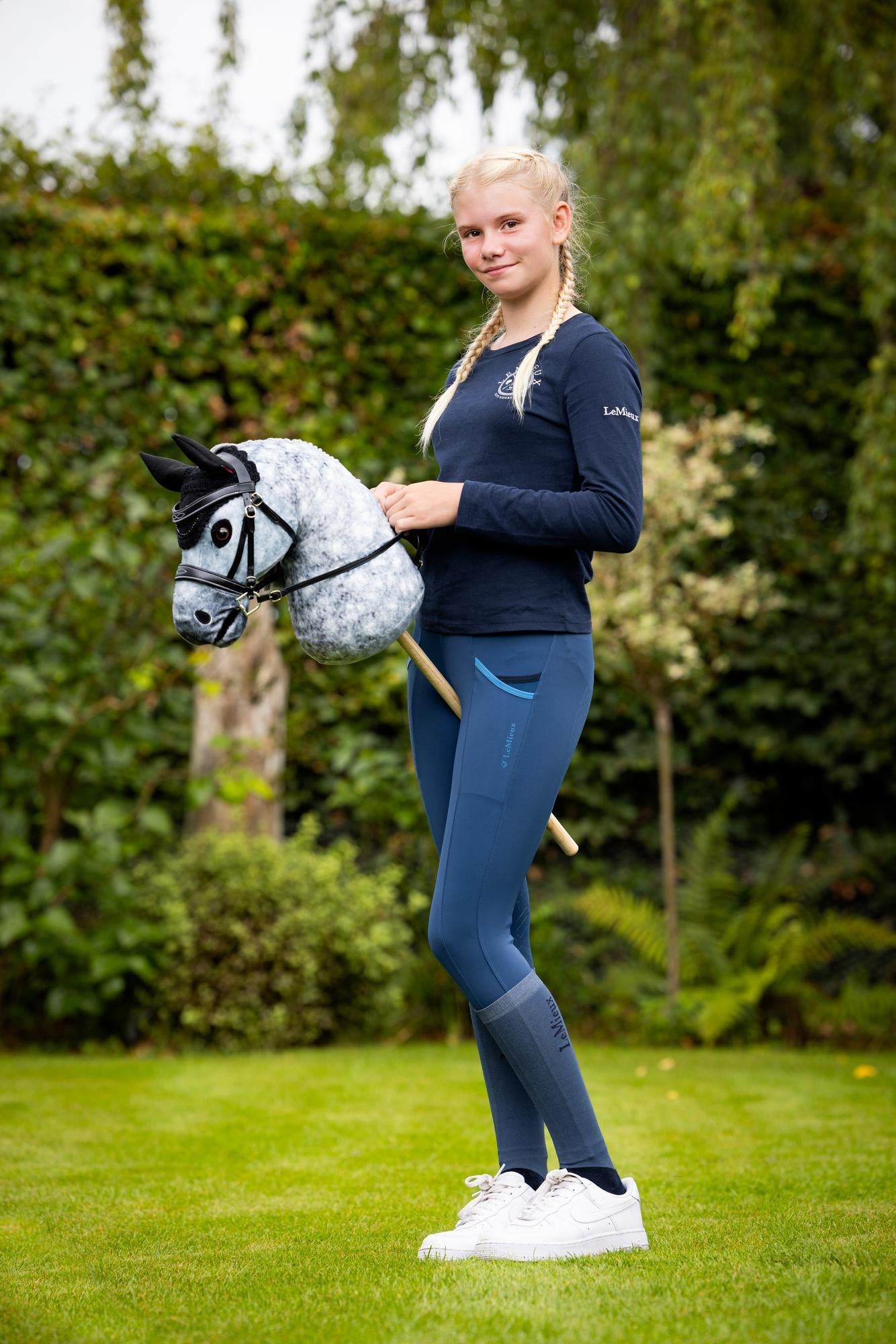 LeMieux Hobby Horse Fliegenmütze Black kombiniert mit dem LeMieux Hobby Horse Sammy und der Hobby Horse Trense Competition und einer Hobby Horse Reiterin im Garten. Zeigt das Zubehör in realistischer Anwendung im Freien, beim Spielen.