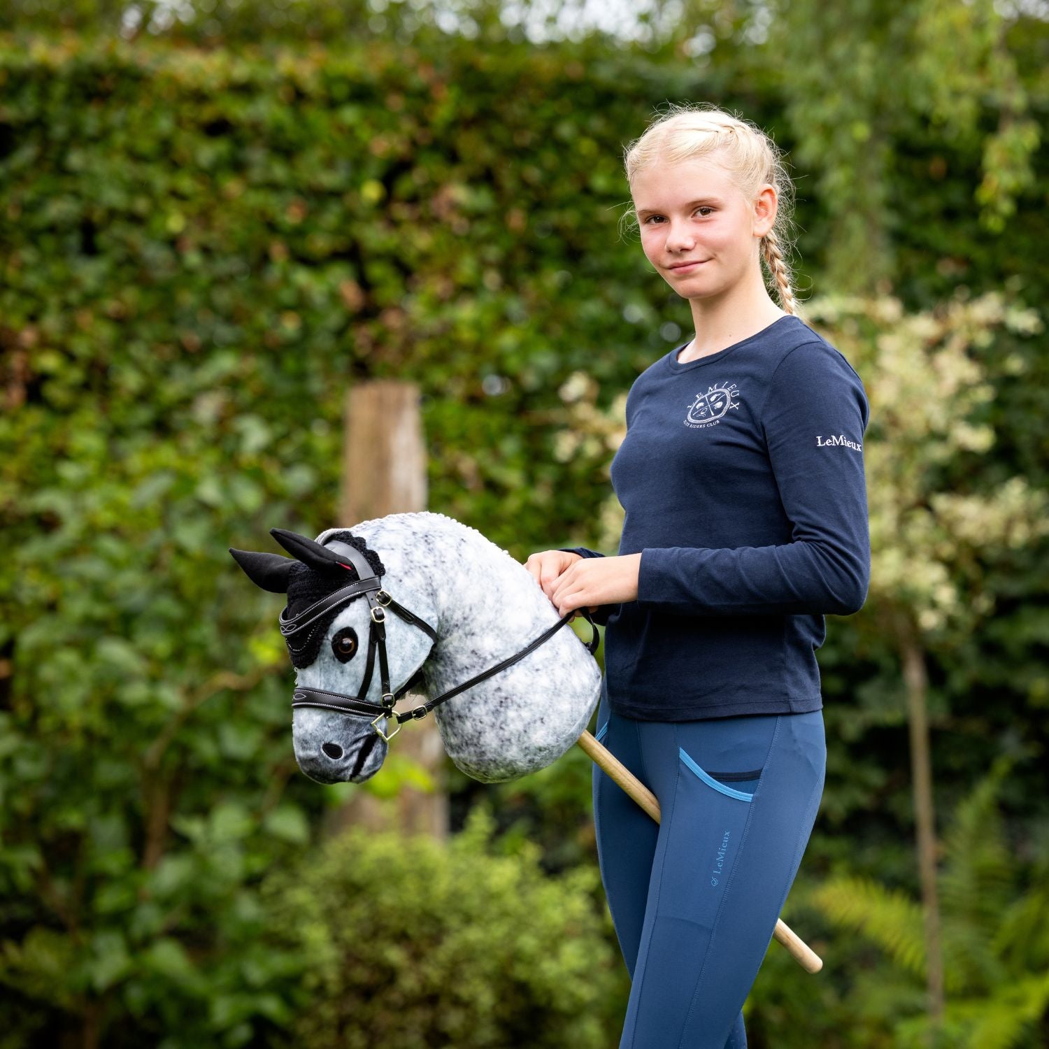 LeMieux Hobby Horse Sammy mit der schwarzen Trense Competition und passender Fliegenmütze mit schwarzen Glitzersteinen. Die junge Hobby Horse Reiterin hält die Zügel in der Hand und trainiert mit Sammy draußen im grünen Garten. Die realistischen Details in Miniaturausführung machen das Hobby Horse und das Hobby Horse Equipment sehr realistisch. Für Kinder und junge Menschen, die Bewegung, Pferde und Spiel lieben.