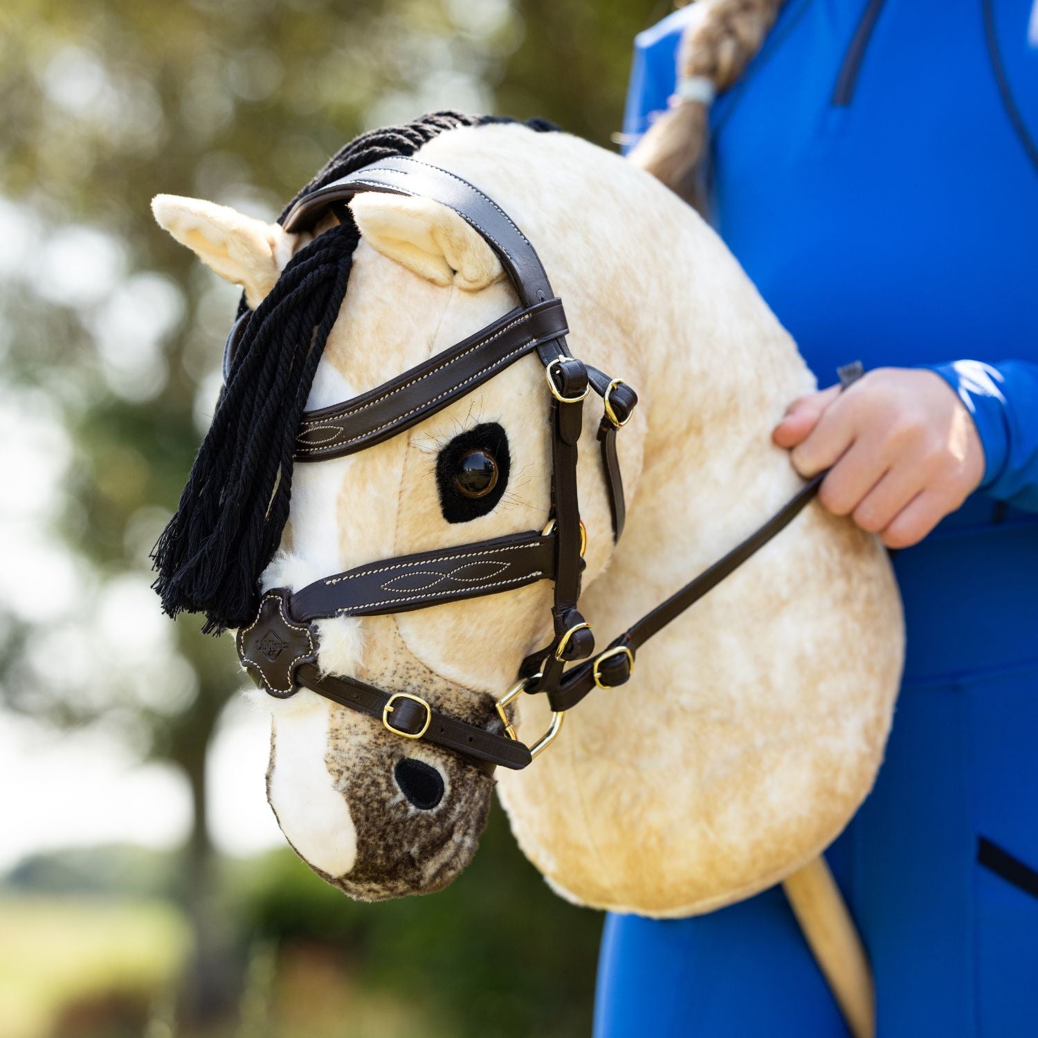 LeMieux Steckenpferd Hobby Horse Dream mit Hobby Horse Trense Grackle, einer mexikanischen Trense zum Springreiten und Turnierreiten. Eine junge Reiterin hält die Zügel der Miniatur Trense mit D-Ring Gebiss fest in der Hand.