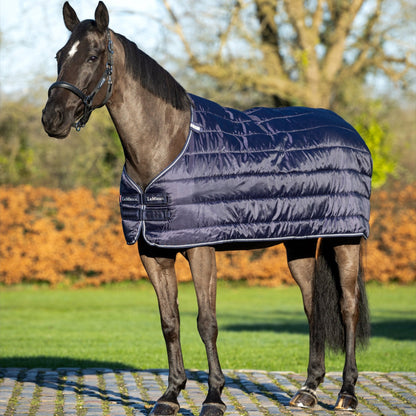 Ein geschorenes Pferd trägt LeMieux Unterdecke 100 g Navy draußen im Sonnenlicht mit herbstlichem Hintergrund leichte seitliche Ansicht glänzende glatte Oberfläche der Decke sichtbarer Brustverschluss und Kassettensteppung elegante dunkelblaue Unterdecke ohne LeMieux Liner-System getragen als Einzeldecke ideal für Wärme und Komfort bei Pferden. 