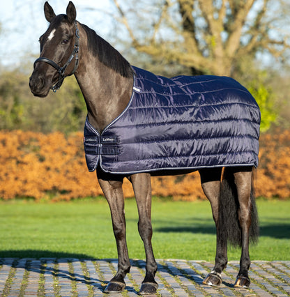Ein braunes Pferd trägt die LeMieux Unterdecke 200 g Navy draußen im Sonnenlicht vor einem herbstlichen Hintergrund. Leichte seitliche Ansicht zeigt die glänzende, glatte Oberfläche der Decke, den sichtbaren Brustverschluss und die Kassettensteppung. Die dunkelblaue Unterdecke wird ohne Liner-System als Einzeldecke getragen und bietet extra Wärme und Komfort.