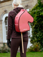 Ein Mädchen trägt die gefüllte LeMieux Hobby Horse Tragetasche Cranberry auf dem Rücken. Durch die Bodenöffnung des Rucksacks schaut der Holzstab des Steckenpferds hervor. Perfekt für unterwegs zum Training oder Turnier.