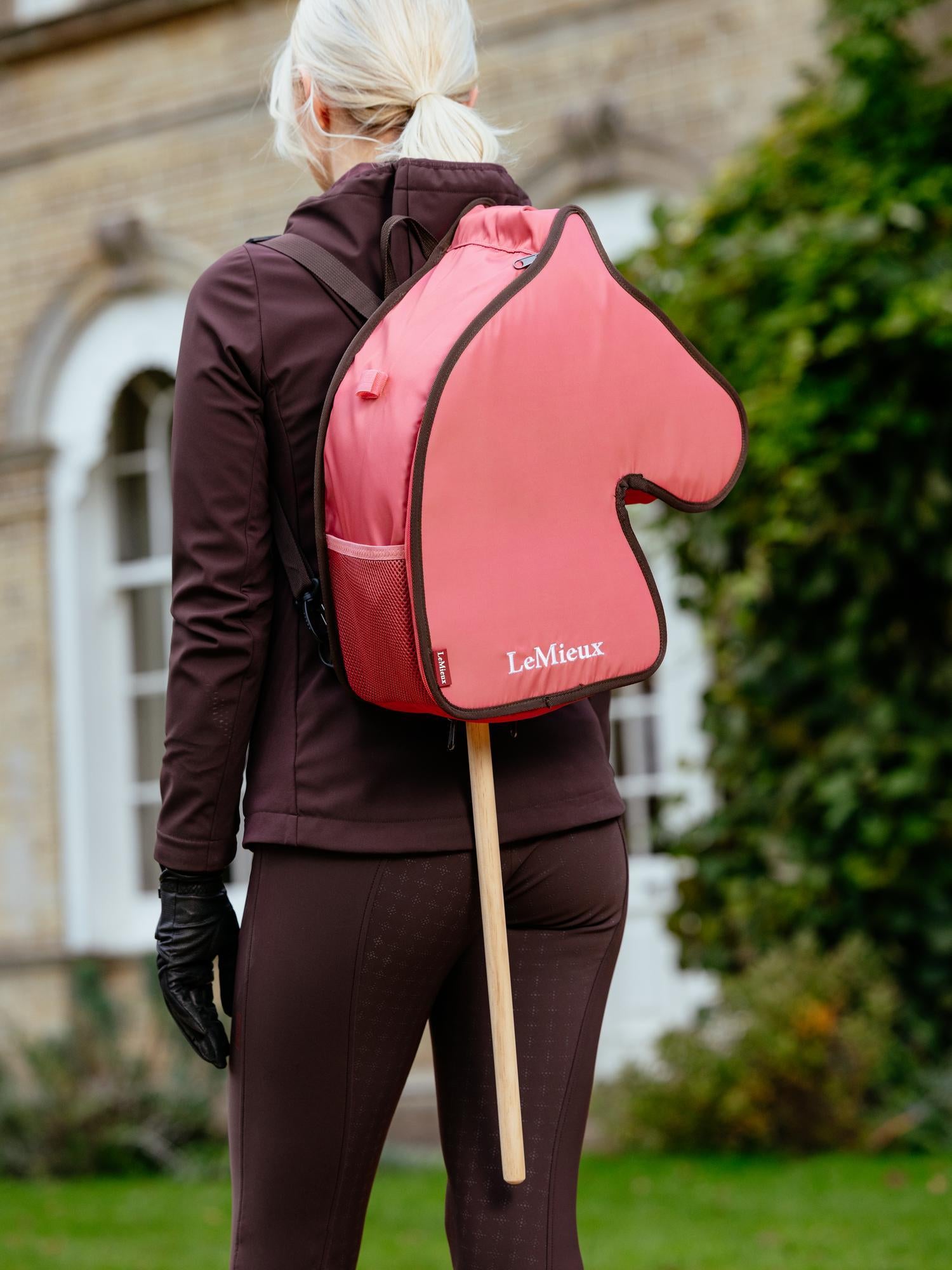Ein Mädchen trägt die gefüllte LeMieux Hobby Horse Tragetasche Cranberry auf dem Rücken. Durch die Bodenöffnung des Rucksacks schaut der Holzstab des Steckenpferds hervor. Perfekt für unterwegs zum Training oder Turnier.