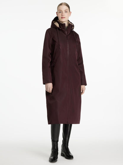 Der lange rotbraune Reitmantel All Elements in der Farbe damson von Lemieux reicht der Reiterin bis zur Wade. Der breite und weite Schnitt wird an der Silhouette der jungen schlanken Frau sichtbar. Der Regenmantel wird geschlossen getragen und lässt viel Raum für freie Bewegungen darin.