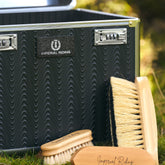 Detailaufnahme der Imperial Riding Putzbox IRHShiny grey black mit geriffelter Wellenstruktur und fühlbarem Relief, schwarz-silbern glitzerndem Imperial Riding Logo sowie sichtbaren Pflegebürsten im Vordergrund – hochwertiges Design einer eleganten Premium Putzbox für Reiterinnen und Reiter.