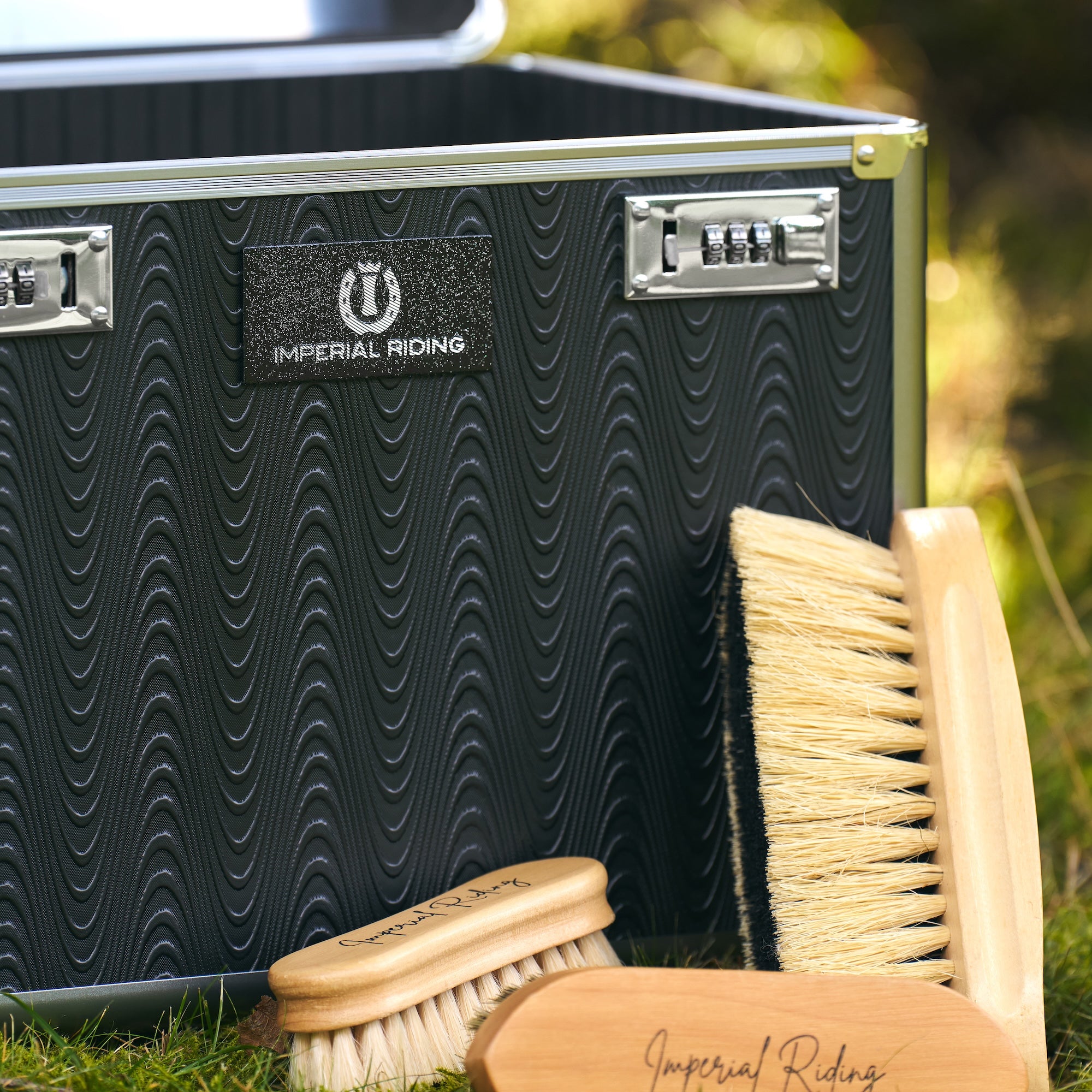 Detailaufnahme der Imperial Riding Putzbox IRHShiny grey black mit geriffelter Wellenstruktur und fühlbarem Relief, schwarz-silbern glitzerndem Imperial Riding Logo sowie sichtbaren Pflegebürsten im Vordergrund – hochwertiges Design einer eleganten Premium Putzbox für Reiterinnen und Reiter.