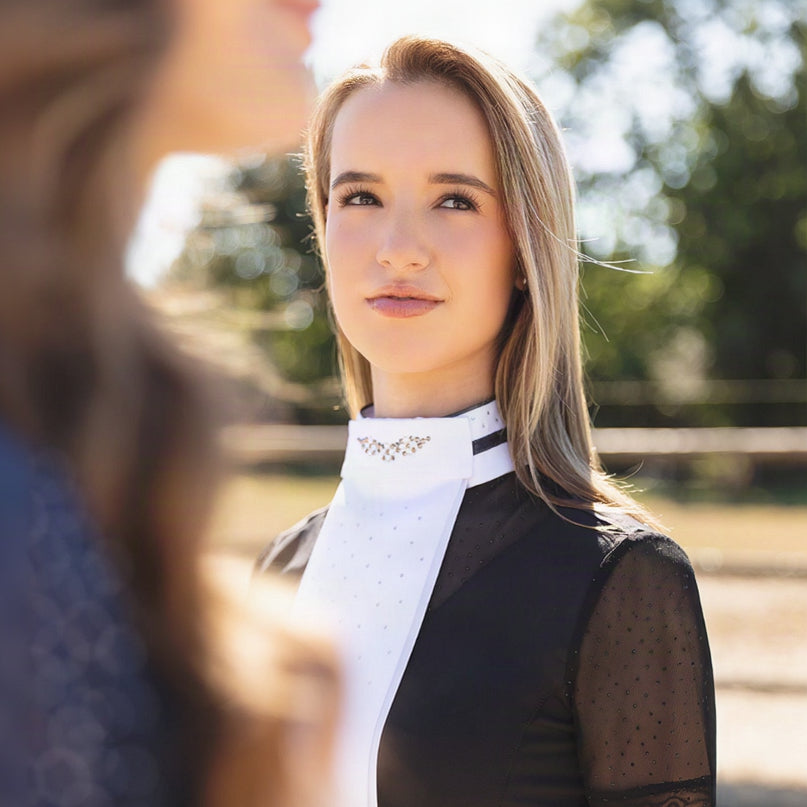 Eine Reiterin im eleganten ANKY Turnieroutfit aus der Competition Wear Collection trägt das ANKY Plastron Prosperous C-Wear White Graphite (ATP26501). In Kombination mit einem dunklen Turniershirt sticht das glitzernde Dressurplastron im Dressurviereck edel hervor.
