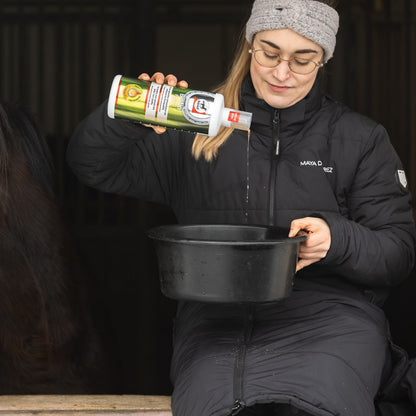 Eine Reiterin mischt das flüssige Zusatzfutter Biotin Plus 3 im Stall unter das Kraftfutter in einem Futtertrog. Sie kippt die zuvor exakt abgemessene Menge aus der Dosierflasche sauber in den Futtereimer. Im Hintergrund ist schemenhaft ihr Pferd in der Box zu erkennen.