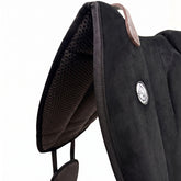 Die Detailaufnahme des schwarzen Brockamp Bareback Pad Reitpad Spezial zeigt das Reitkissen von vorne mit Blick auf die strukturierte, rutschfeste Anti-Slip-Unterseite aus Neopren im Übergang zur feinen Wildleder-Oberfläche. Erkennbar sind das runde Brockamp-Logo-Emblem, der braune Ledergriff mit Kontrastnaht und die robuste Kantenverarbeitung für optimalen Halt beim Reiten ohne Sattel.