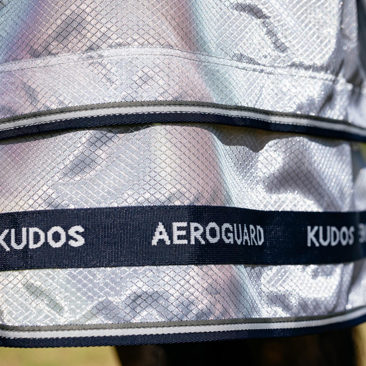 Glänzendes Detailbild des hochtechnischen Materials der LeMieux Insektenschutzdecke in Silber. Gut sichtbar ist der spezielle, zweiteilige Saum im Bereich der Hinterbeine für einen perfekten Fall der Decke, stilvoll abgerundet durch die Kudos AeroGuard Logo-Aufschrift auf den robusten, dunkelblauen Einfassungen.