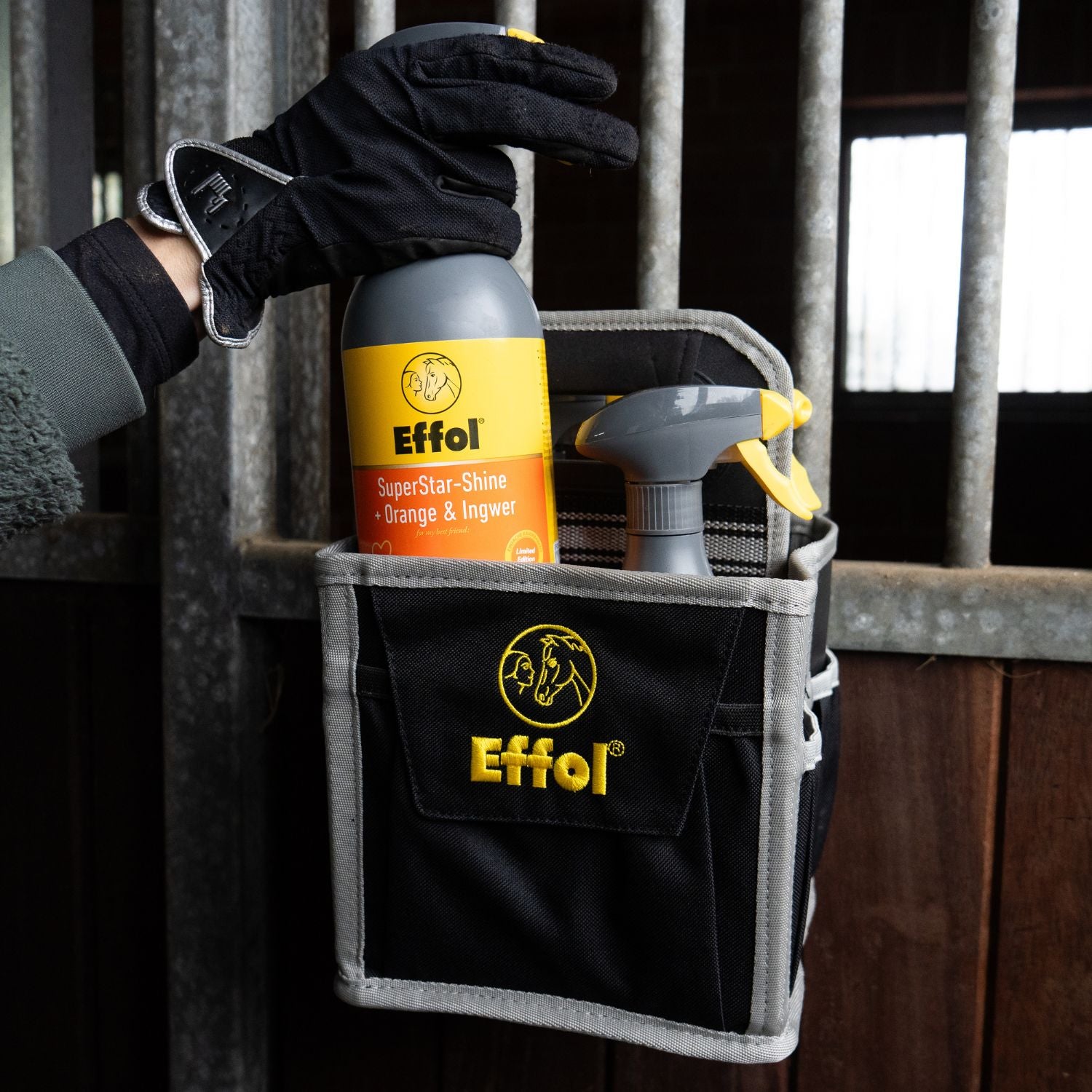 Die kleine offene Putztasche hängt an der Boxentür und ist gefüllt mit großen 750-ml-Sprühflaschen von Effol. Eine der Flaschen wird gerade herausgezogen und soll verwendet werden. Dabei handelt es sich um das Schweif- und Mähnenspray „SuperStar Shine Orange-Ingwer“ mit orangem Etikett. Eine absolute Besonderheit unter allen Glanzsprays im Reitsport und in der Pferdepflege.
