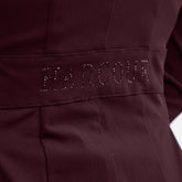 Seitliches Detailbild der Rückseite des Harcour Turnierjackets Oria in Aubergine. Im Bereich der Taille betont ein angedeuteter Bund den femininen Schnitt der Turnierjacke für Damen. Ein funkelnder Harcour-Logo-Schriftzug aus großen, auberginefarbenen Strasssteinen ziert diesen Taillenbund. Dieses wunderschöne Glitzerdetail wertet die dezente Rückansicht auf und macht das Reitsakko für Markenfans zu einem echten Designerstück, das die weibliche Silhouette im Turniersport elegant in Szene setzt.
